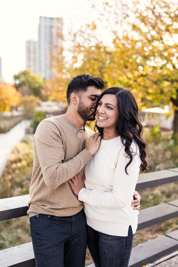 Man speaks in his partner's ear as they stand close together, smiling, on a Lincoln Park bridge with autumn trees and city buildings in the background during their Chicago fall engagement session