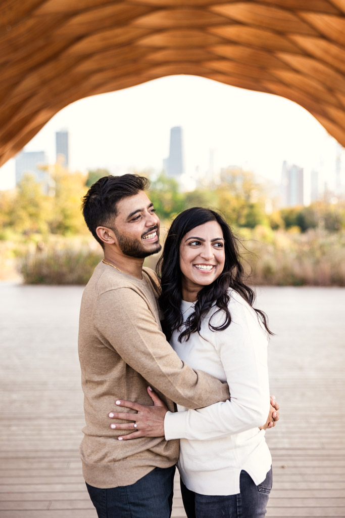 A smiling couple holds each other under the Lincoln Park Honeycomb with the Chicago skyline in the background during their fall engagement session