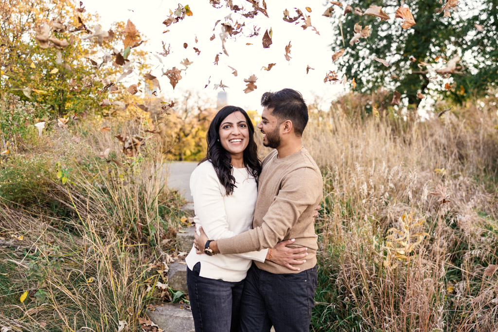 A couple embraces on a Lincoln Park path in autumn during their Chicago fall engagement session, smiling happily as leaves swirl around them