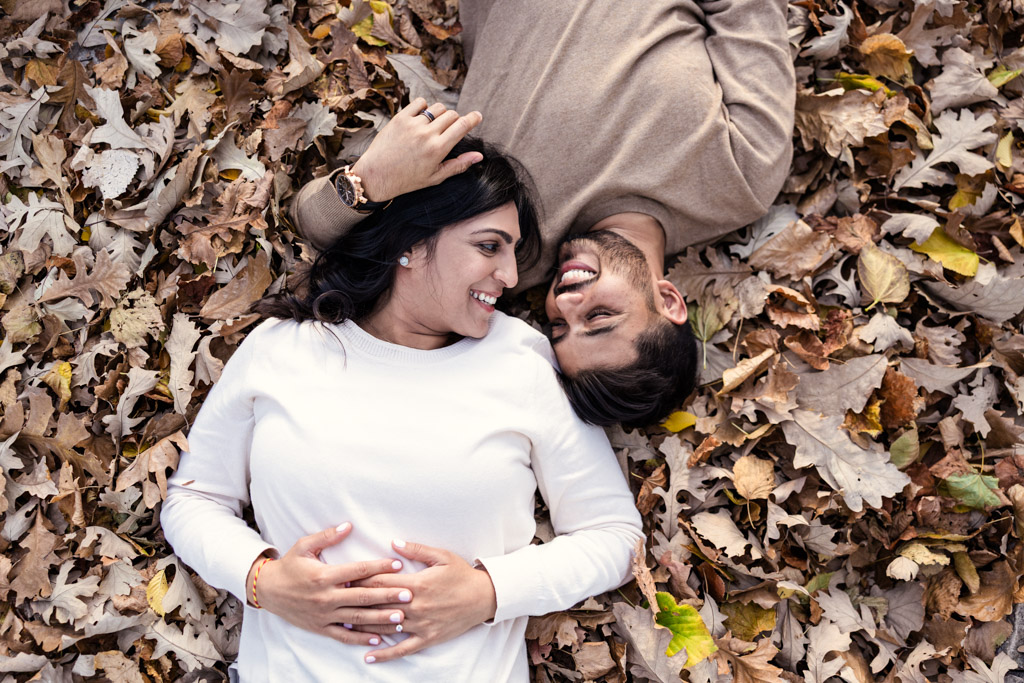 A smiling couple lies on autumn leaves, facing each other and holding hands during their Chicago fall engagement.