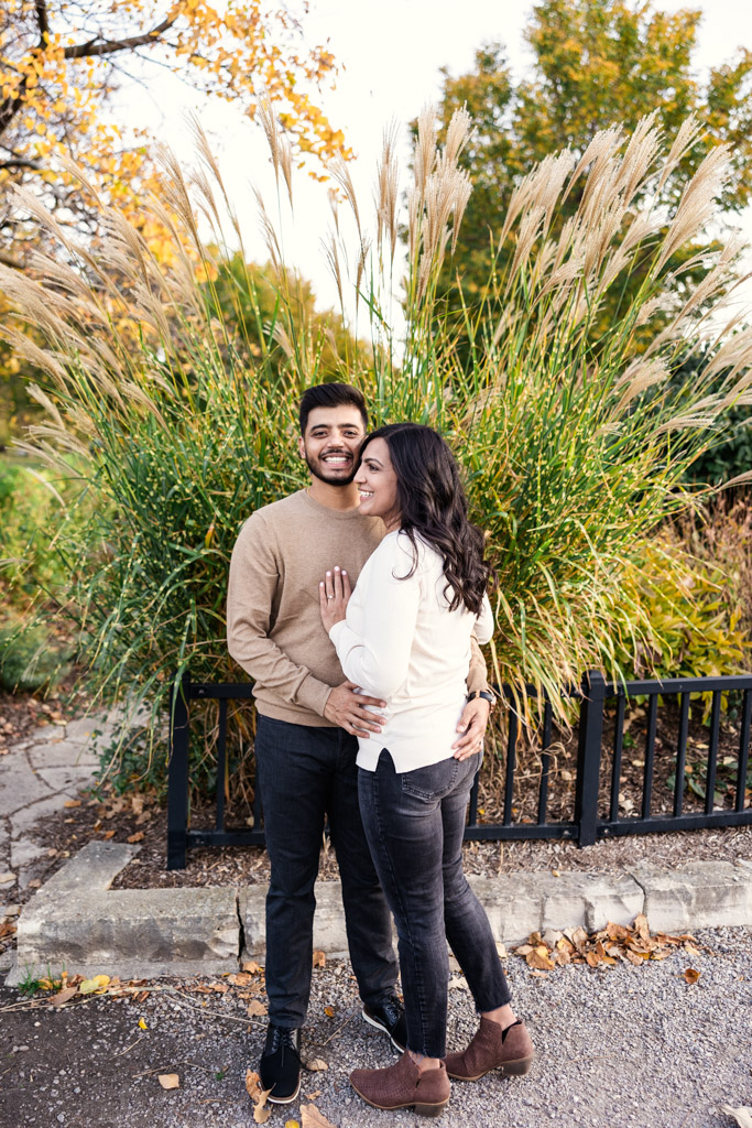 Happy engaged couple stands close together, smiling, with tall grass and autumn trees behind them in Lincoln Park