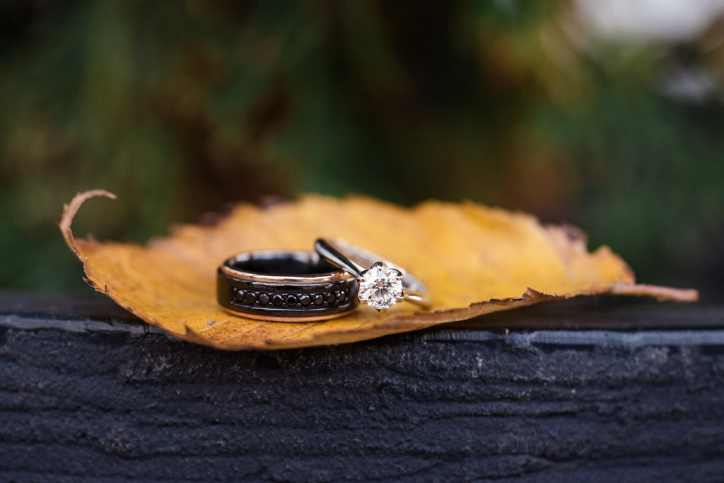 Two rings, one with a diamond, rest on a yellow leaf atop a dark wooden surface
