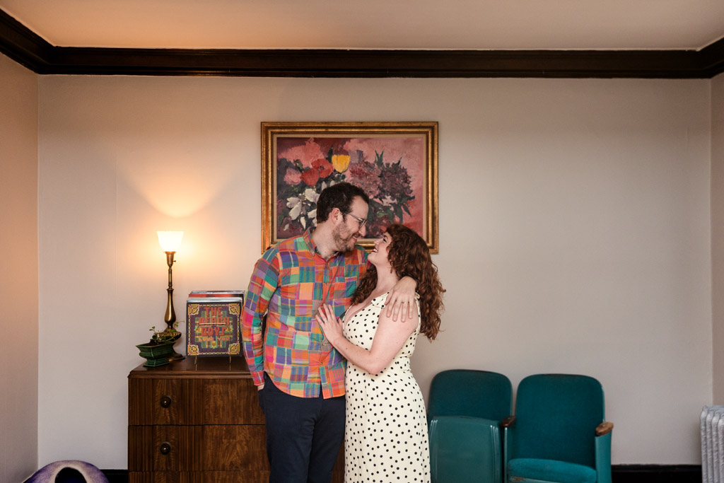 A couple stands close together and smiles in a living room during their Chicago home engagement session, with a floral painting on the wall behind them