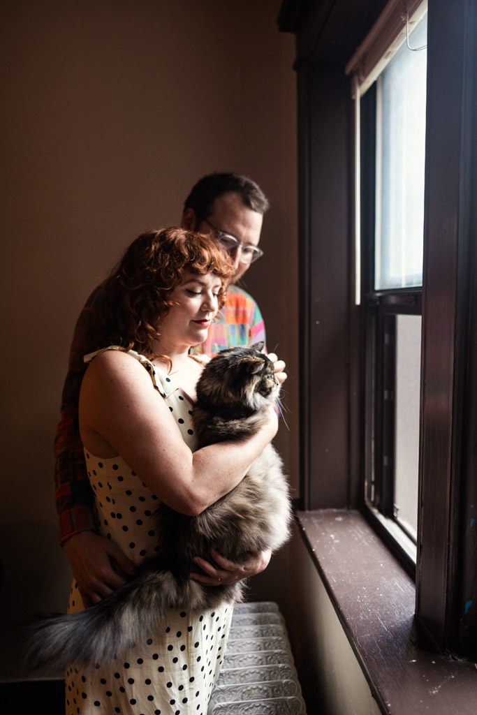 A woman holds a cat stands by a window in her Chicago home, with her partner hugging her from behind