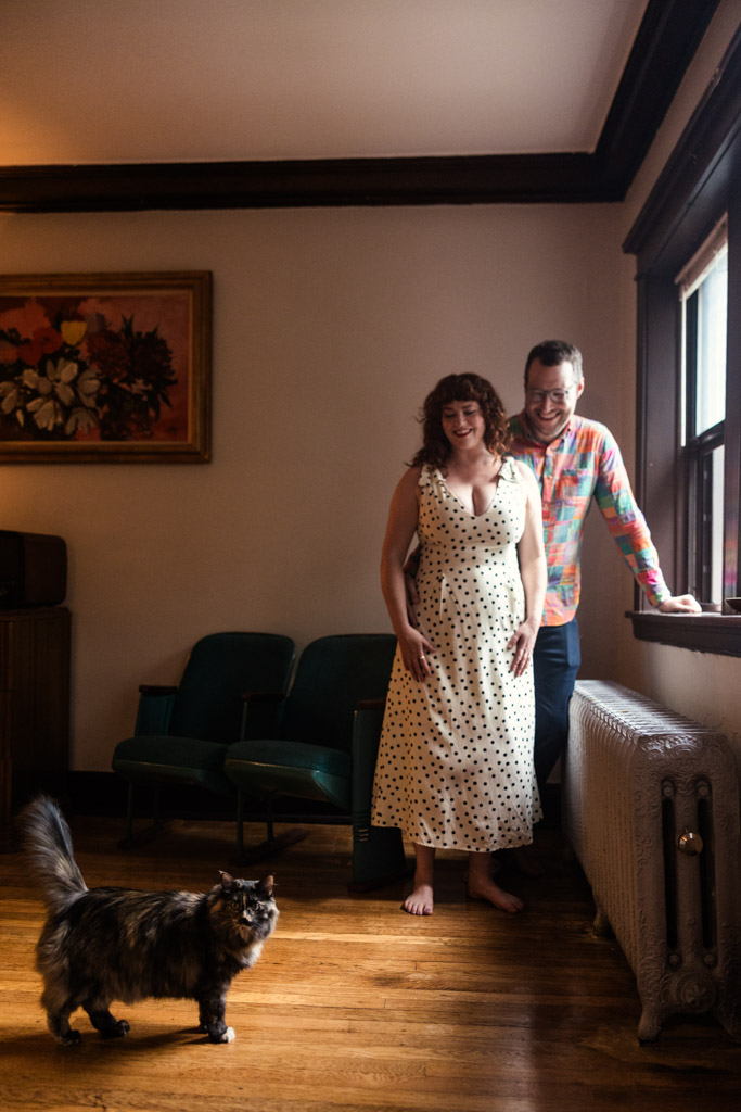 Happy engaged couple stands by a window in their living room watching their cat walk across the wooden floor during their Chciago home engagement session