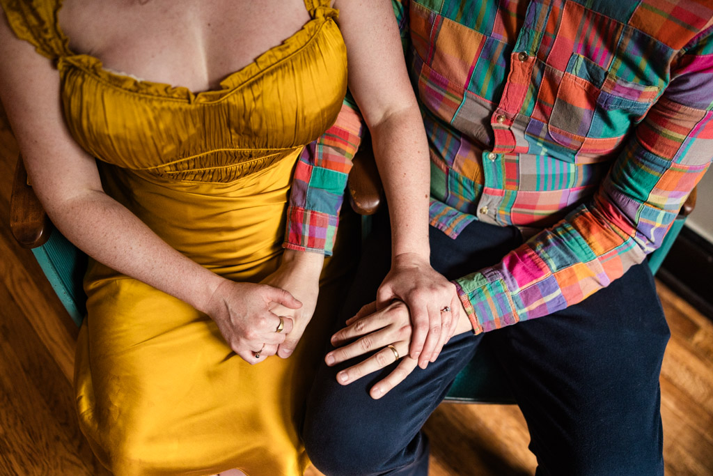 A couple holding hands—one in a yellow dress, the other in a colorful plaid shirt—sit together during their cozy Chicago home engagement session