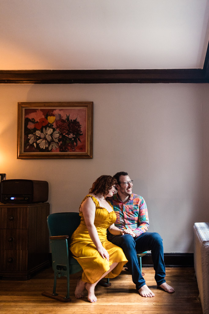 Engaged couple sits closely together on a chair in their sunlit Chicago home, smiling, with artwork hanging on the wall behind them