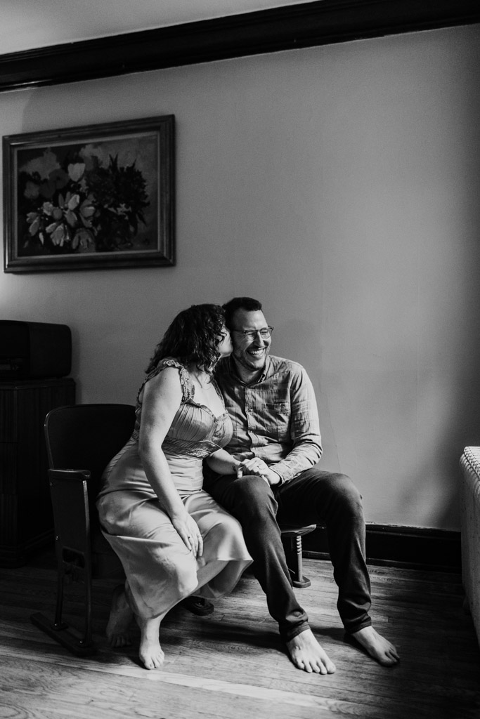 Black and white photo of woman kissing her partner on the cheek as they sit barefoot together in their Ravenswood home during Chicago engagement session