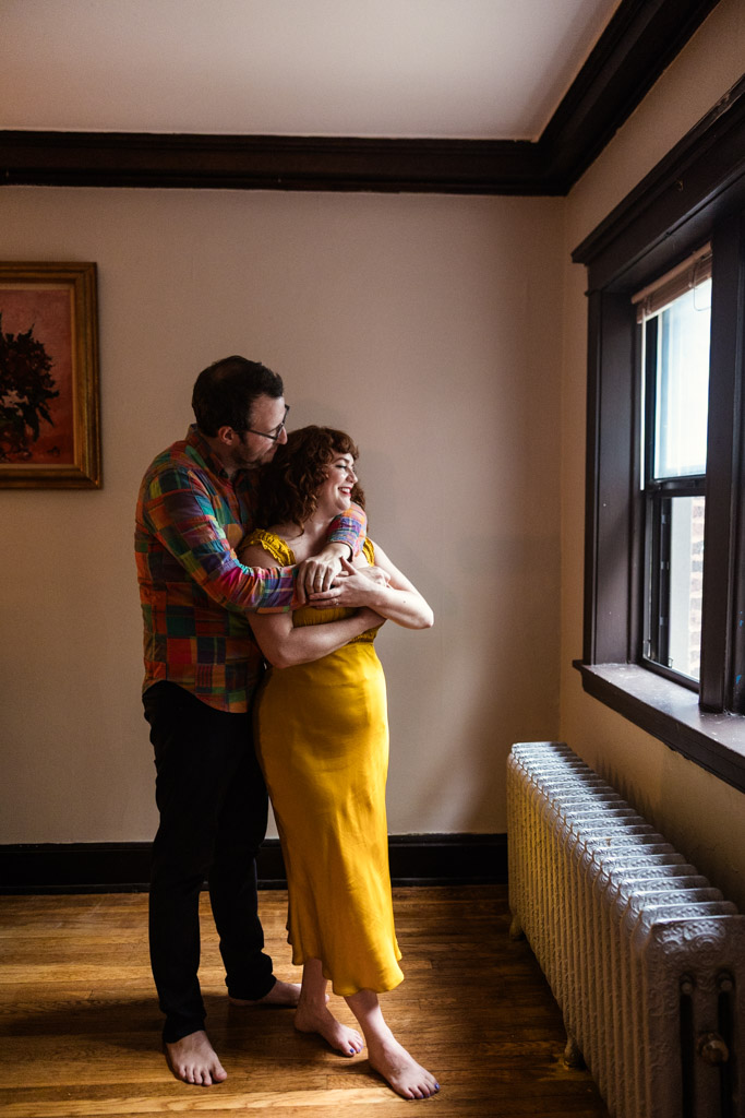 Happy couple stands barefoot by a window, embracing and smiling in a warmly lit room during their cozy Chicago home engagement session