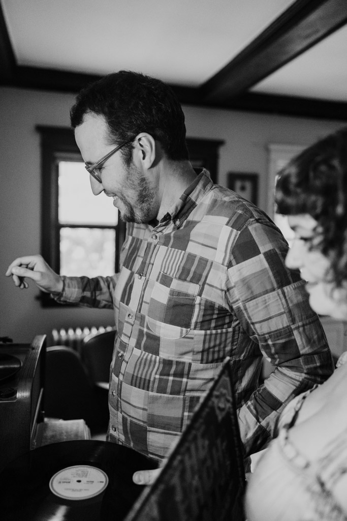 Black and white photo of a man in glasses and a plaid shirt playing a vinyl record while his partner stands beside him