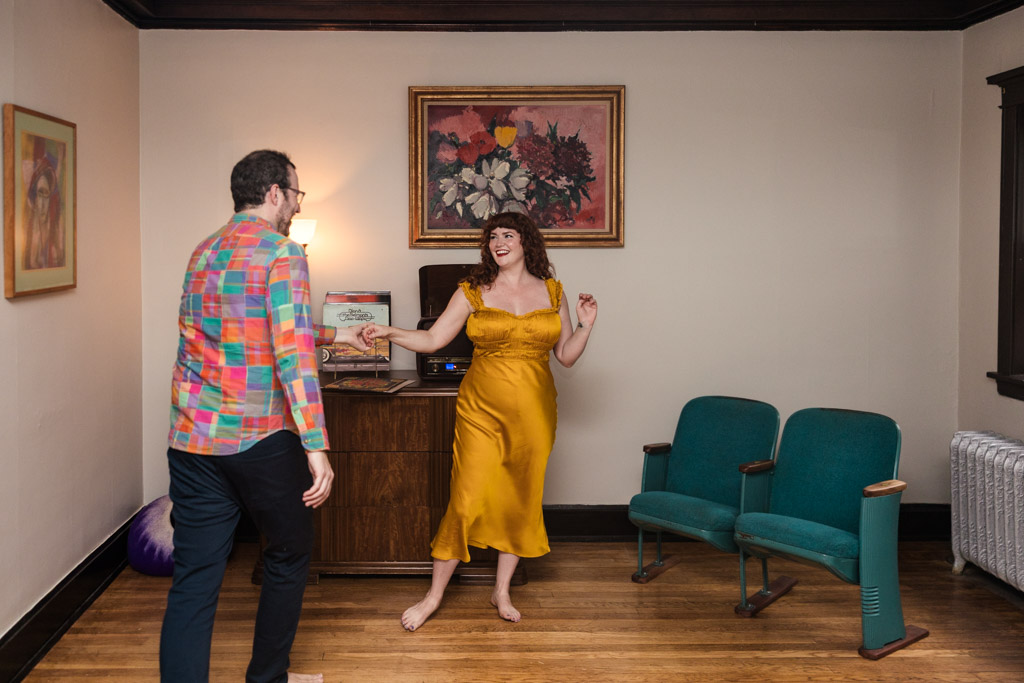 Happy engaged couple dances barefoot in their Chicago home. The woman wears a yellow dress and the man a colorful shirt