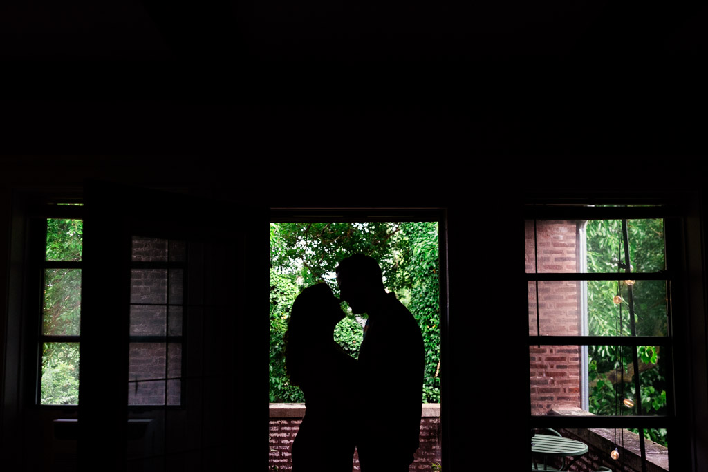 Creative photo of couple facing each other silhouetted in front of a window in their home, with greenery and brick visible outside during their Chicago home engagement session