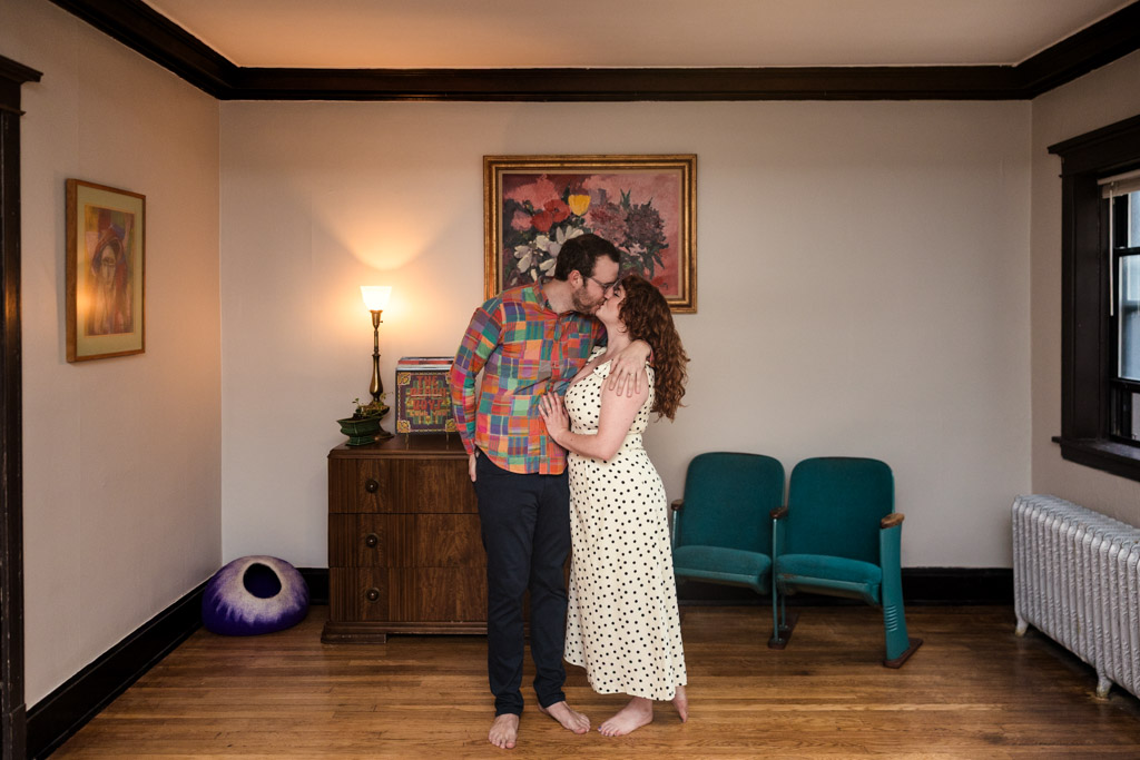 Engaged couple stands barefoot, embracing and kissing in the living room of their Chicago home
