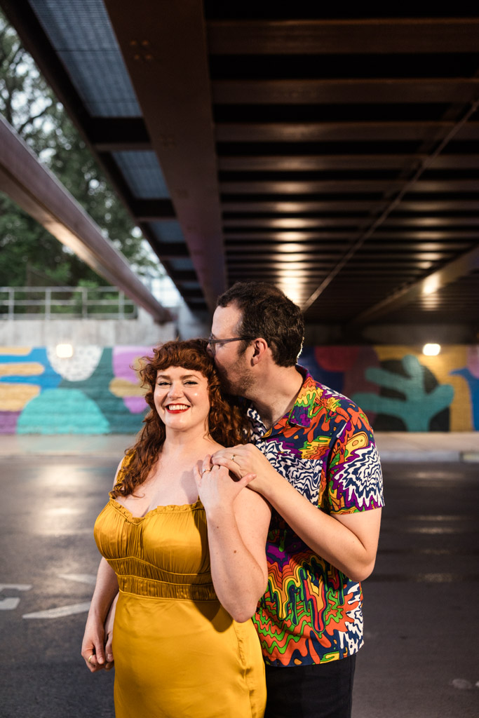 Happy woman in a yellow dress stands with her partner wearing a colorful shirt under a bridge with mural art during Chicago engagement sessioon