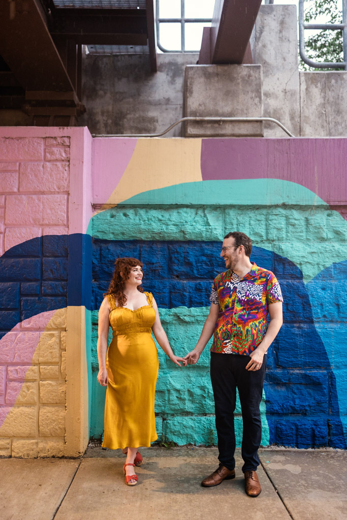 Engaged couple holding hands and smiling, standing in front of a colorful mural under a train overpass in Ravenswood