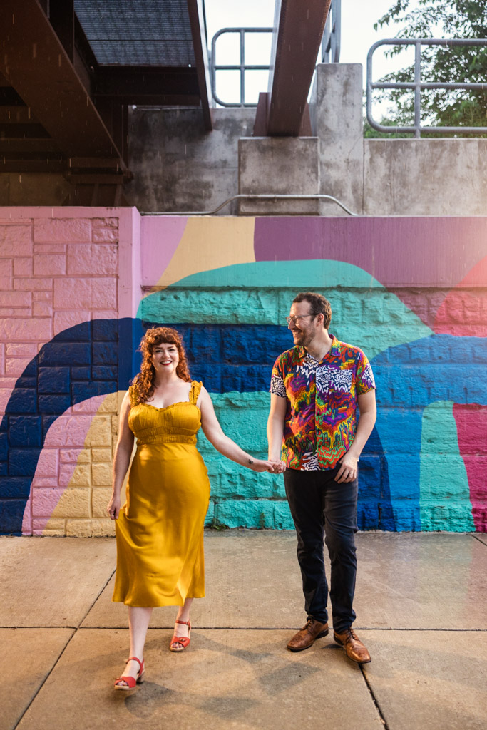Happy couple holding hands walks in front of a colorful mural under an overpass in the Ravenswood neighborhood of Chicago during their, the woman wearing a yellow dress and the man wearing a colorful shirt