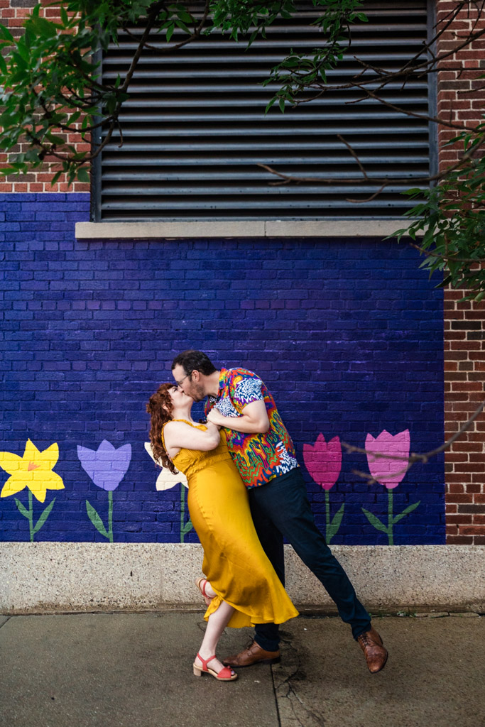 Engaged couple kisses in front of a purple brick wall painted with colorful flowers while walking through Ravenswood Chicago home engagement.
