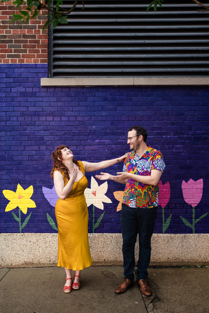 A woman in a yellow dress and a man in a colorful shirt laugh by a flower mural on a purple brick wall during their Chicago engagement session