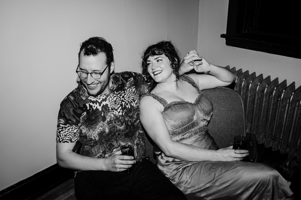 Black and white photo of engaged couple sitting close, smiling, holding drinks, in their Ravenswood home