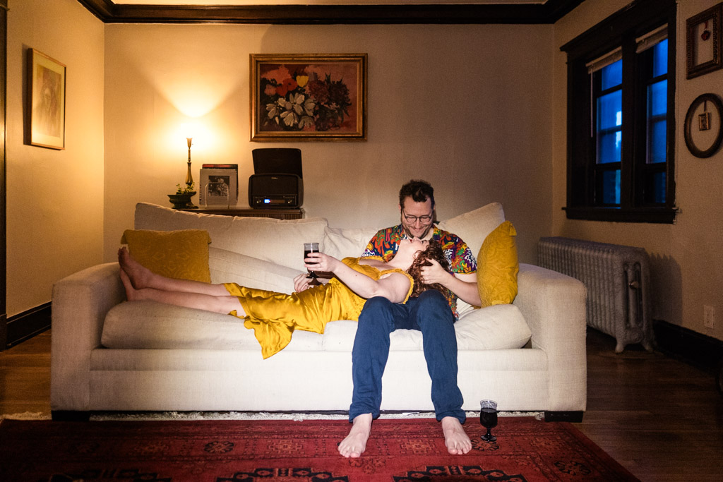 A couple relaxes on a couch in their Chicago home; the woman lays across the man’s lap, both holding wine glasses during their cozy Chicago engagement session