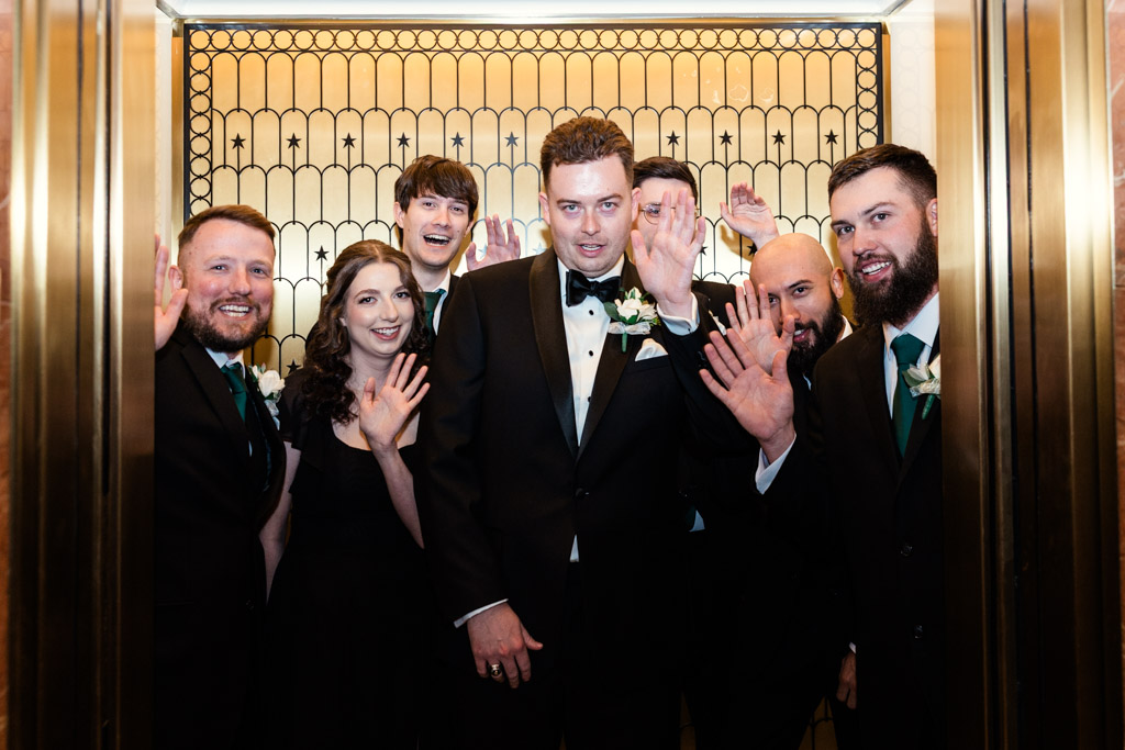 Groom and groomspeople in formal wear smiling and waving inside a golden elevator before Library at 190 wedding ceremony
