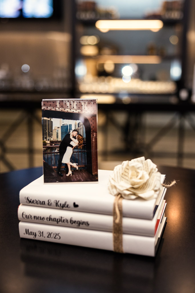 Custom decor piece with photo of couple kissing on top of stacked books labeled with their names and wedding date, beside a paper flower at The Library at 190 in downtown Chicago