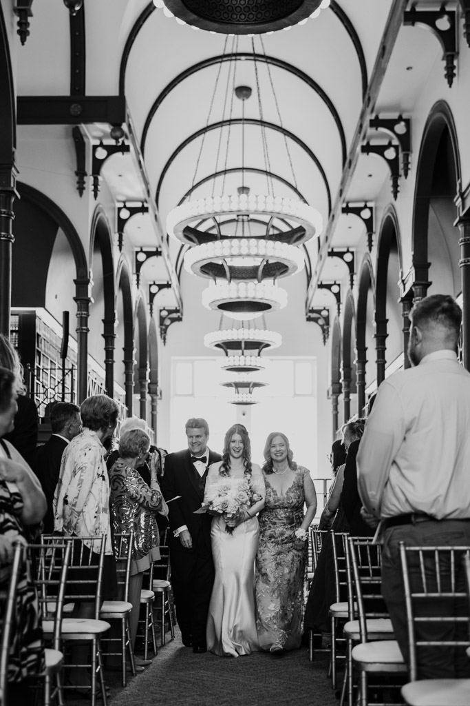 Black and white photo of bride walking down the aisle with her parents, surrounded by seated guests in wedding ceremony space at The Library at 190 in downtown Chicago