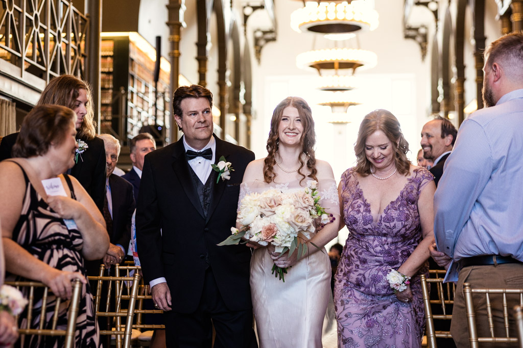 Bride in white dress walks down the aisle with her smiling parents during wedding ceremony at The Library at 190 in downtown Chicago