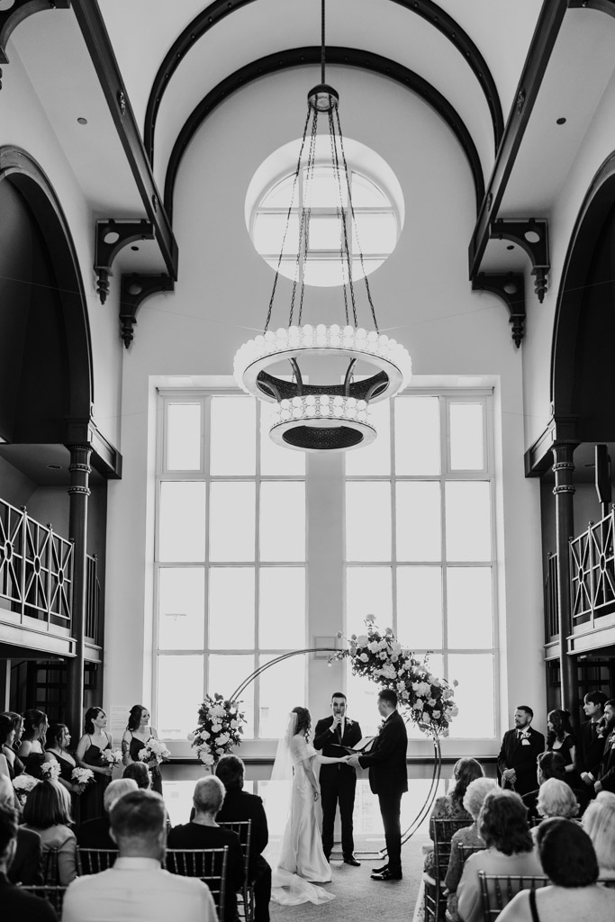 Black and white photo of groom placing ring on bride's finger with a large arched window and chandelier, during wedding ceremony at The Library at 190 in downtown Chicago