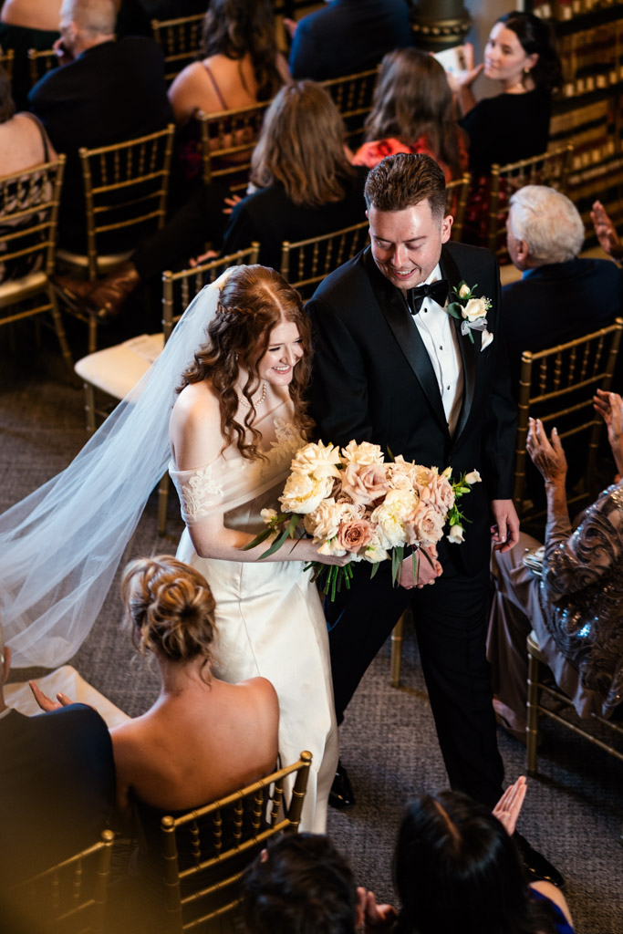 Bride and groom smiling and walking down the aisle surrounded by seated guests after their Library at 190 wedding ceremony