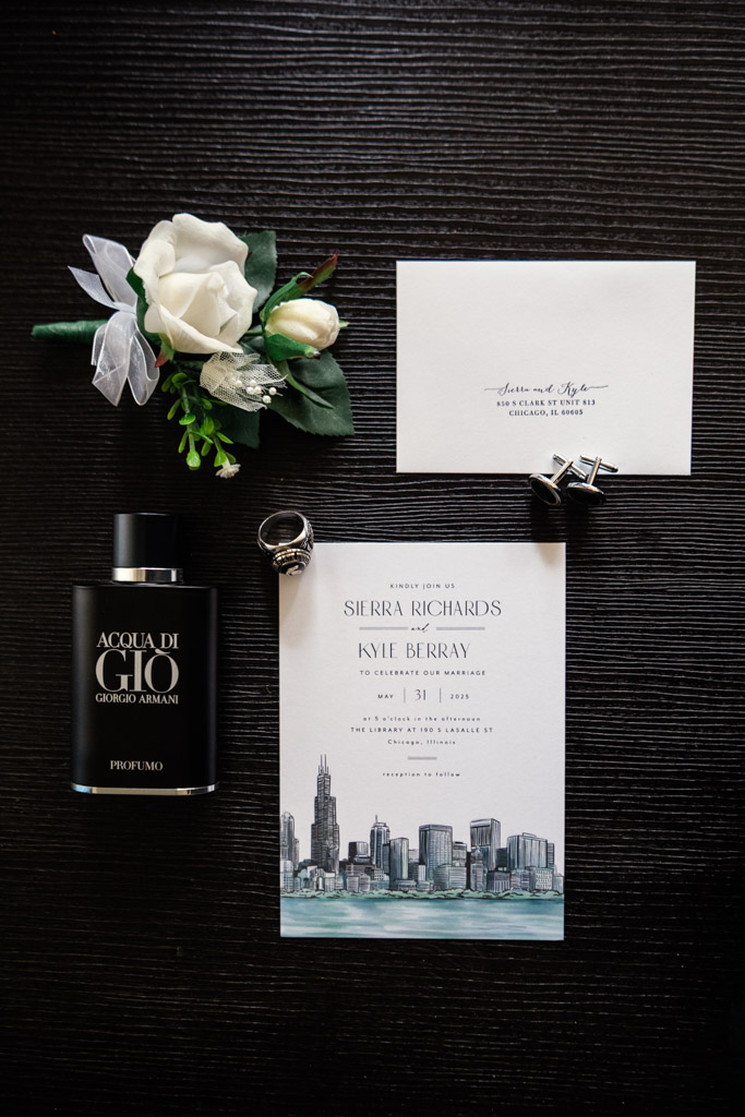 Flat lay of wedding invitation, rings, perfume, cufflinks, boutonniere, and envelope on a black surface for wedding at The Library at 190 in downtown Chicago