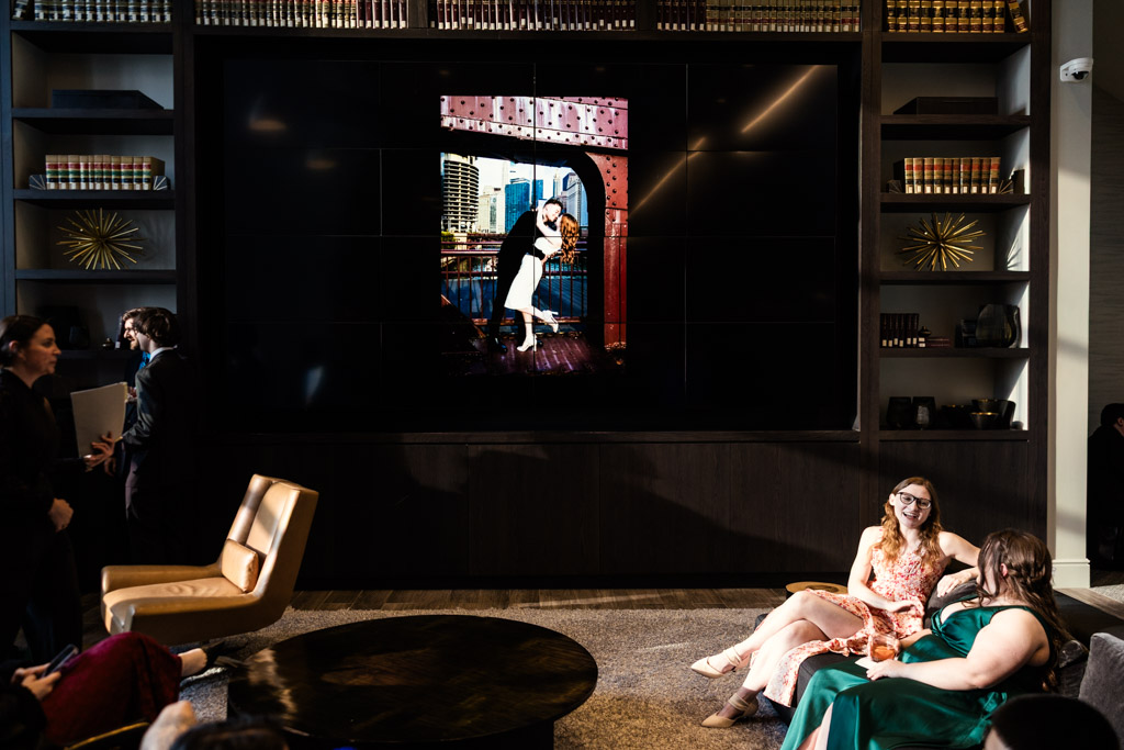 Two guests in formal dresses sit on a couchduring cocktail hour at The Library at 190 in downtown Chicago; a large photo of the bride and groom kissing is displayed behind them