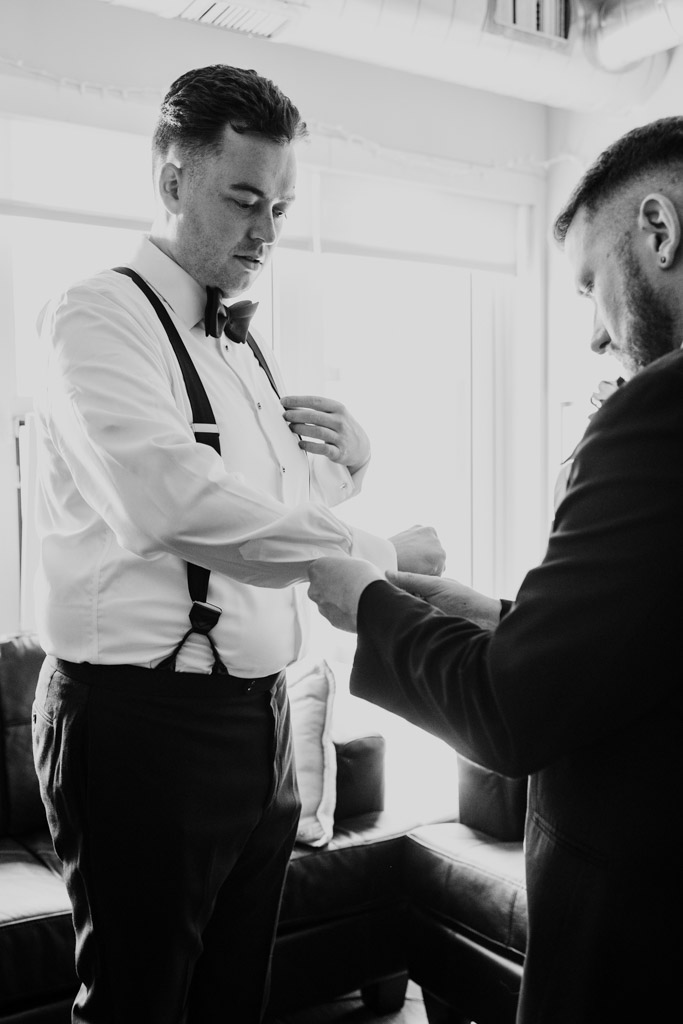 Black and white photo of Best Man helping groom with cufflinks in a bright room before wedding at The Library at 190 in downtown Chicago