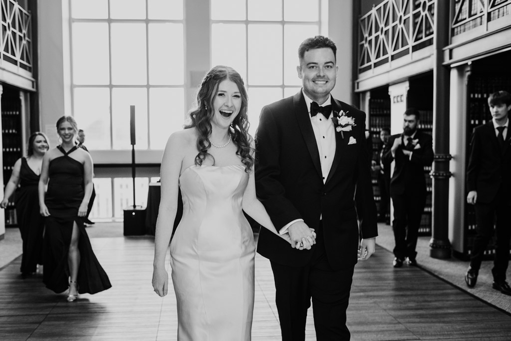 Black and white photo of bride and groom walking hand in hand, smiling, with their wedding party following behind them at their Library at 190 wedding reception