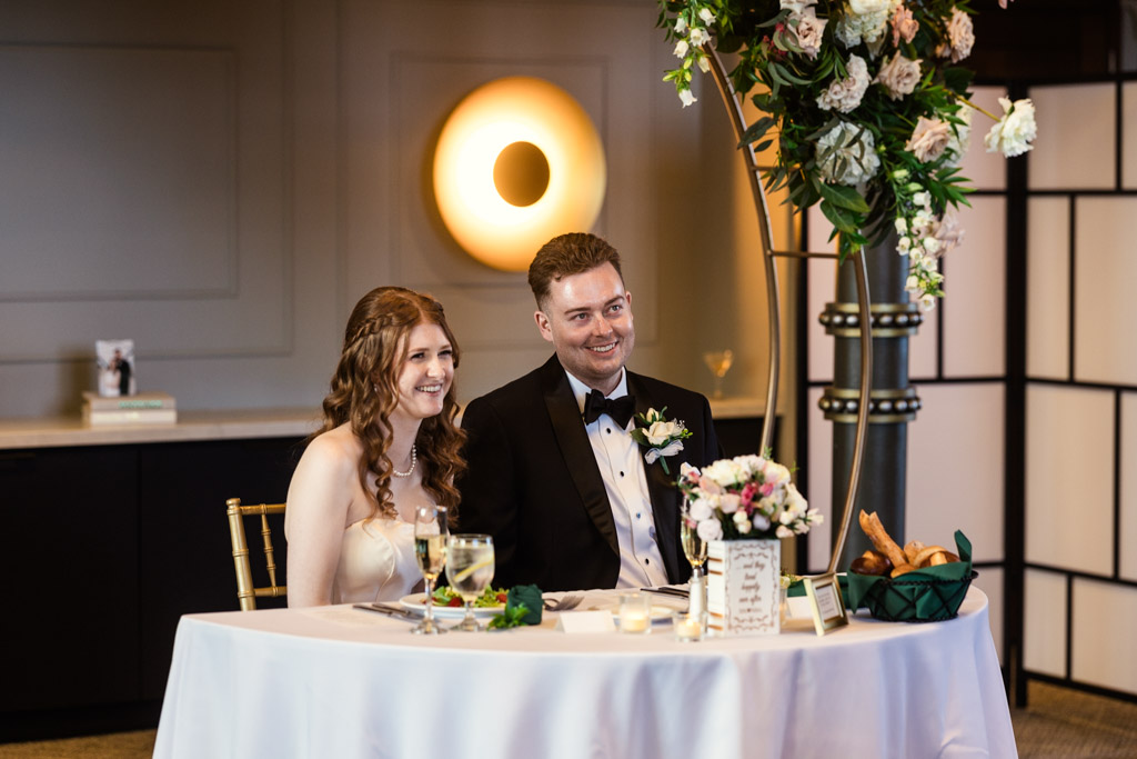 Bride and groom sit smiling at a decorated table listening to a speech during their The Library at 190 wedding reception