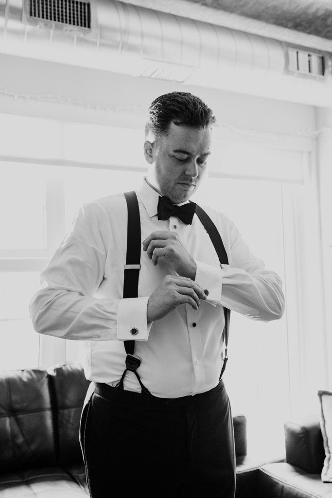 Ggroom adjusts his cufflink, wearing suspenders and a bow tie in a bright room before his wedding at The Library at 190 in downtown Chicago