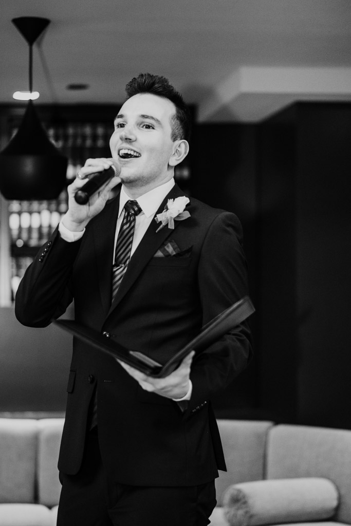 Black and white photo of officiant in a suit holding a microphone and speaking during wedding reception at The Library at 190 in downtown Chicago