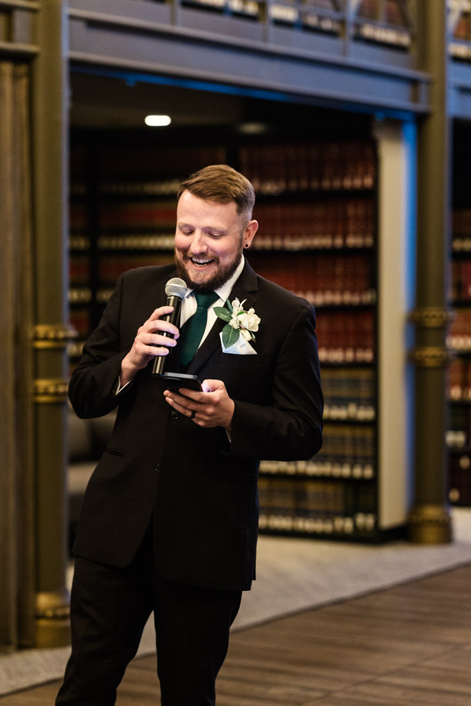 Best Man in a suit with a boutonniere speaks into a microphone, smiling and reading from his phone during wedding reception at The Library at 190 in downtown Chicago