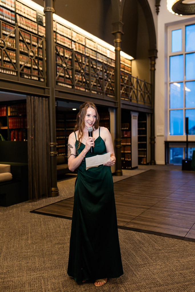 Maid of Honor in a green dress gives speech with tall bookshelves in the background during wedding reception at The Library at 190 in downtown Chicago