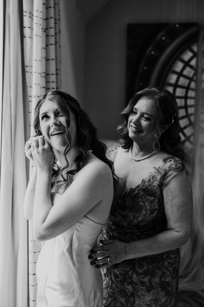 Bride smiles while putting on earrings as her mother helps her with her dress before her wedding at The Library at 190 in downtown Chicago