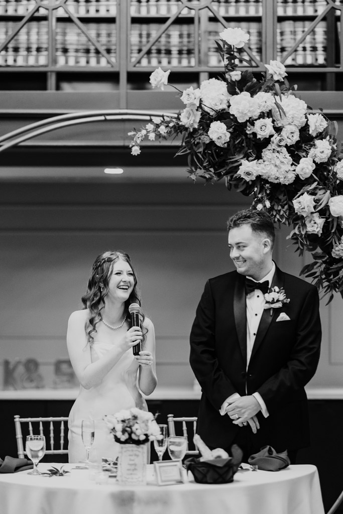 Black and white photo of bride laughng while giving a speech next to smiling groom beneath a floral arch during their wedding reception at The Library at 190 in downtown Chicago