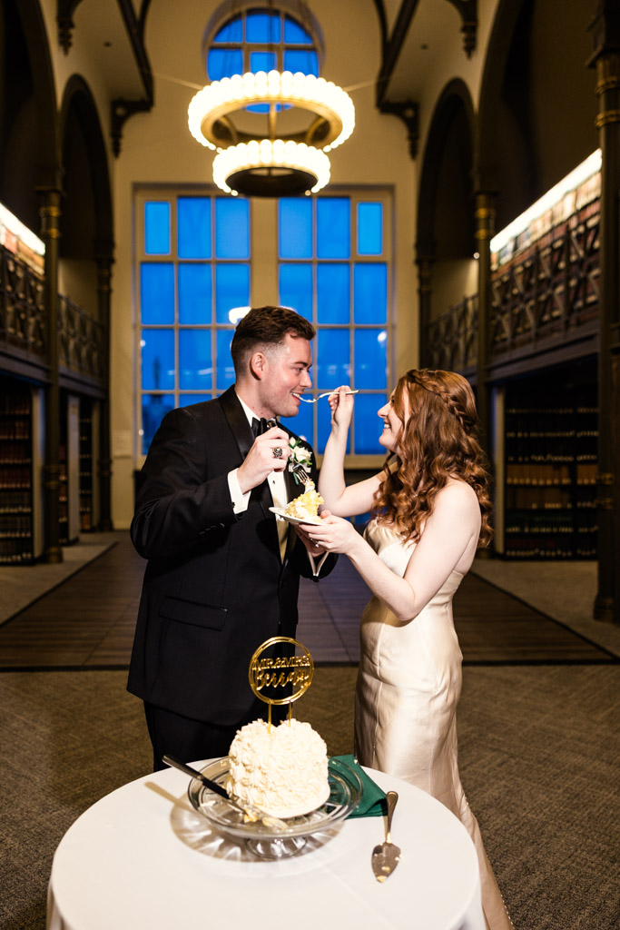 Bride and groom feed each other cake in an elegant room with high ceilings and large windows during their Library at 190 wedding reception