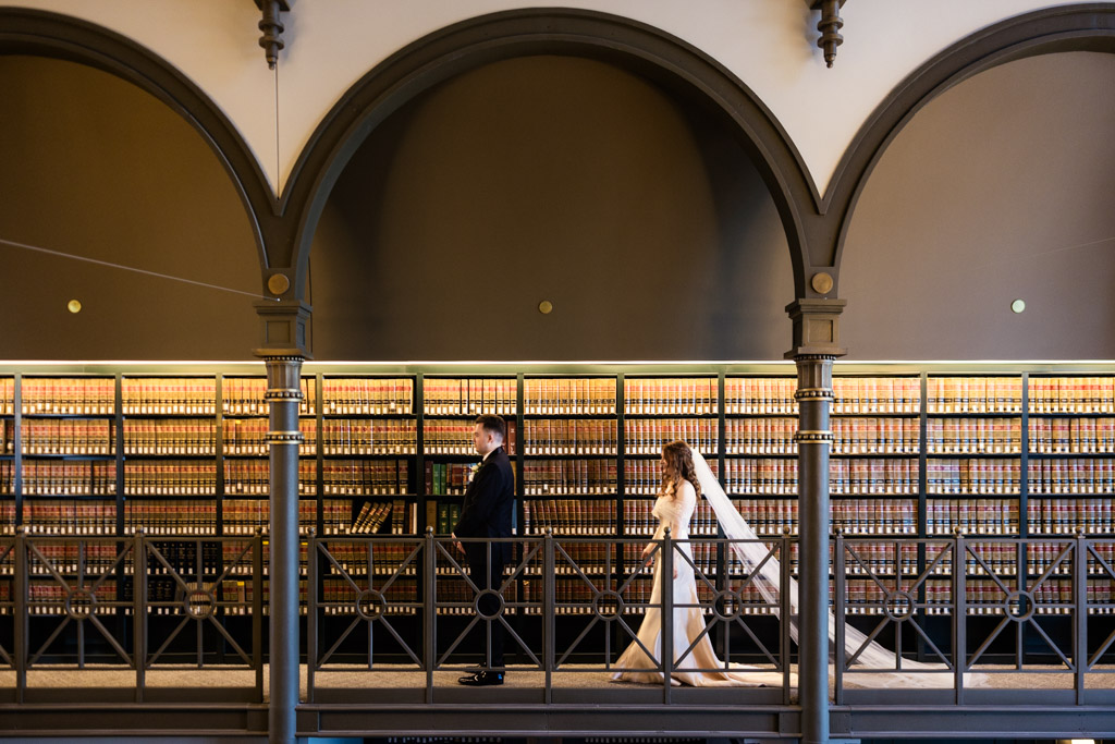 Bride approaches groom on a balcony at The Library at 190 during their first look, shelves of books behind them