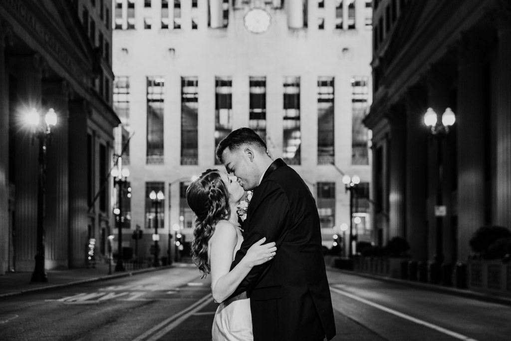 Black and white photo of newlywed couple kissing in the middle of an empty city street at night, surrounded by tall buildings, outside The Library at 190 in downtown Chicago