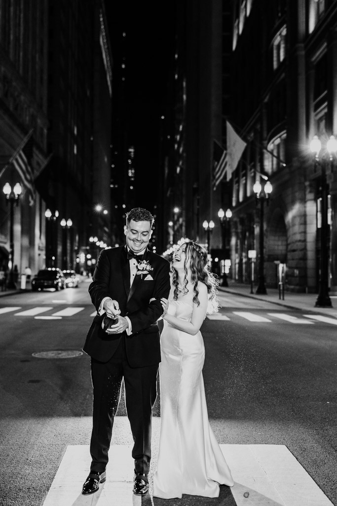 Black and white photo of happy bride and groom popping champagne on LaSalle Street at night outside The Library at 190 in downtown Chicago