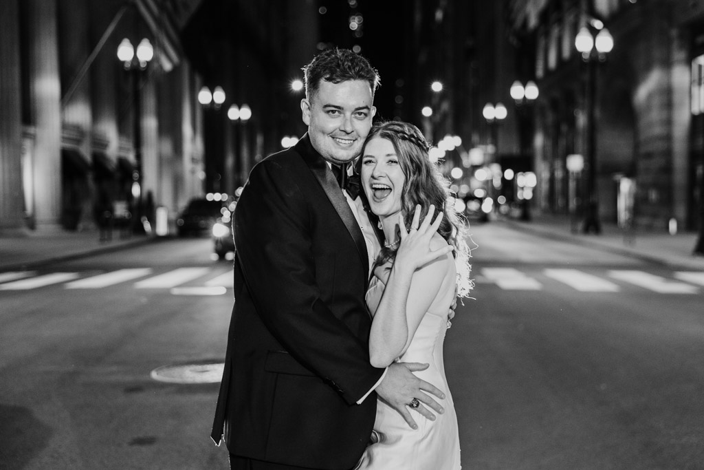 Black and white photo of happy newlywed couple posing on LaSalle Street in downtown Chicago at night with bride displaying her ring during their Library at 190 wedding reception