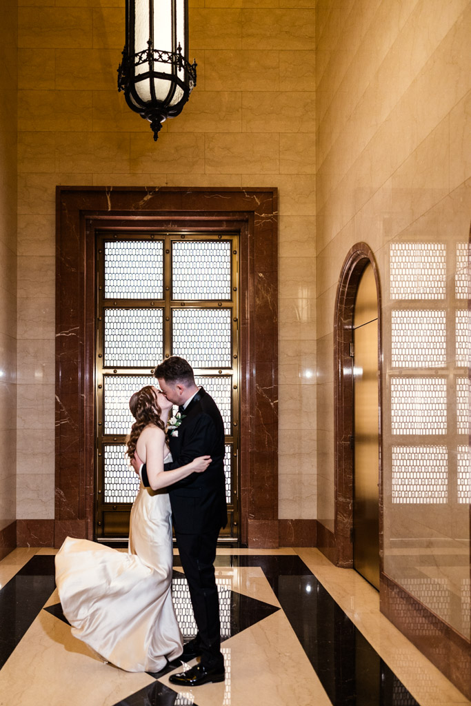 Bride and groom kiss in an, marble-lined hallway under a large chandelier during their wedding celebration at The Library at 190 in downtown Chicago