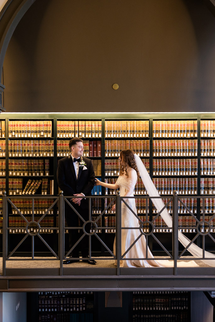 Bride and groom stand on a balcony in front of shelves filled with books, smiling at each other during their first look at The Library at 190