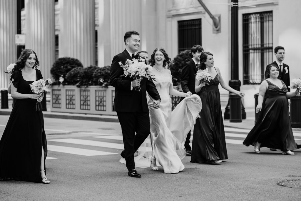 Black and white photo of wedding party walking down LaSalle Street near The Library at 190 in downtown Chicago smiling and holding bouquets, dressed in formal attire