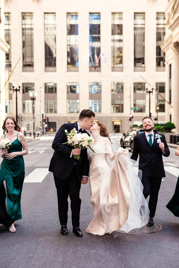 Bride and groom kiss while walking with their wedding party on LaSalle Street before their wedding at The Library at 190 in downtown Chicago